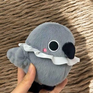 Könnte beinhalten: Ein grauer Plüschvogel mit einem weißen Rüschenkragen, einem schwarzen Schnabel und großen schwarzen Augen mit weißen Umrissen. Der Vogel hat eine kleine rosa Wange und einen kleinen Flügel. Es ist ein Stofftier.