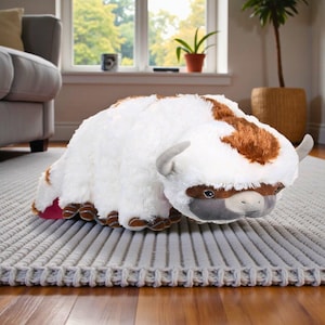 Pode incluir: Um brinquedo de pelúcia em forma de um bisonte branco e marrom, com chifres cinzentos e rosto cinzento. O brinquedo está sobre um tapete cinzento, num chão de madeira. O bisonte tem manchas marrons na cabeça e nas costas.