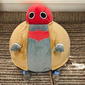May include: A plush toy resembling the DC Comics character Red Hood. The toy features a red helmet with white-rimmed eyes, a tan coat, and a gray shirt with a red bat symbol. The toy has small arms and legs and is sitting on a brown rug.