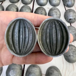 May include: Two antique, silver-colored metal molds with a ribbed, oval design are held in a hand. The molds are open, revealing their interior. Several other similar molds are scattered in the background, suggesting a collection or set.