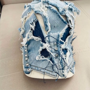 Pode incluir: Boné de beisebol de ganga desgastada com um design de patchwork. O boné apresenta vários tons de ganga azul, com bordas desfiadas e detalhes de tachas prateadas ao longo da aba. O boné tem uma parte inferior branca.