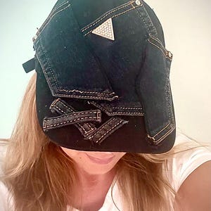 Op de afbeelding: Een zwarte honkbalpet met donkere denim patches op de voorpanelen gestikt. De pet bevat een driehoekig metalen embleem met kleine ingebedde kristallen en verschillende gerafelde denim stroken die over het onderste voorste gedeelte liggen.