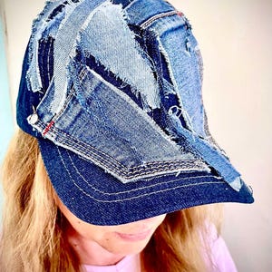 May include: A baseball cap constructed from multiple overlapping patches of blue denim fabric. The design features frayed edges, visible stitching, and varying shades of indigo and light blue denim, creating a textured, layered appearance.