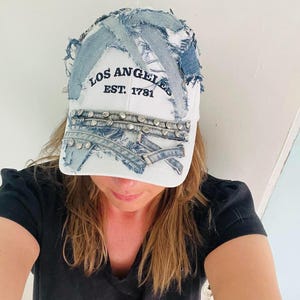 May include: A white baseball cap featuring distressed blue denim fabric patches layered across the crown and brim. The cap displays the black embroidered text LOS ANGELES EST. 1781 and is accented with rows of silver-toned metal studs on the brim.