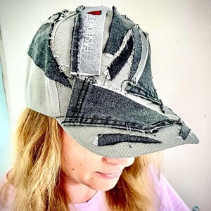 Può includere: Un cappellino da baseball grigio con toppe in denim grigio scuro dall'aspetto consumato e bordi sfilacciati. Una striscia di tessuto rettangolare sulla corona riporta la parola FASHION in lettere bianche con un piccolo triangolo rosso.