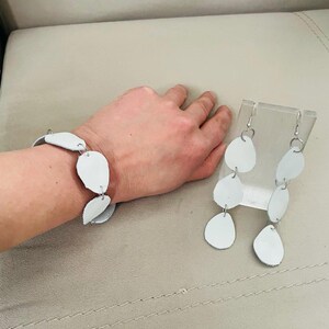 May include: White leather bracelet and matching earrings. The bracelet features multiple white, oval-shaped leather pieces connected by silver links. The earrings have a similar design, with three white leather ovals suspended from silver hooks. The jewelry is displayed on a neutral background.