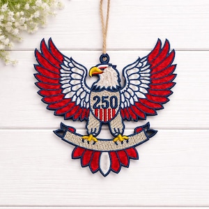 May include: Embroidered eagle ornament with red, white, and blue details. The eagle has outstretched wings and a banner below. The number "250" is displayed on the eagle's chest. A jute string is attached for hanging.