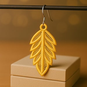 May include: A single yellow leaf-shaped earring with a silver hook. The earring is made of embroidered fabric, featuring intricate details and a delicate, openwork design. The earring is hanging against a blurred background.