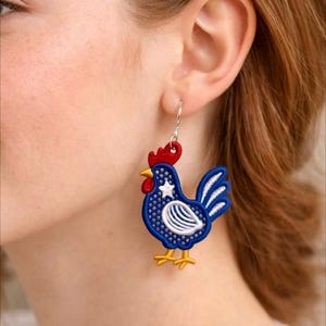 May include: A pair of rooster-shaped earrings. The earrings feature a blue body with white and red accents, including a white star. The rooster has a red comb and yellow feet. The earrings are hanging from silver hooks.