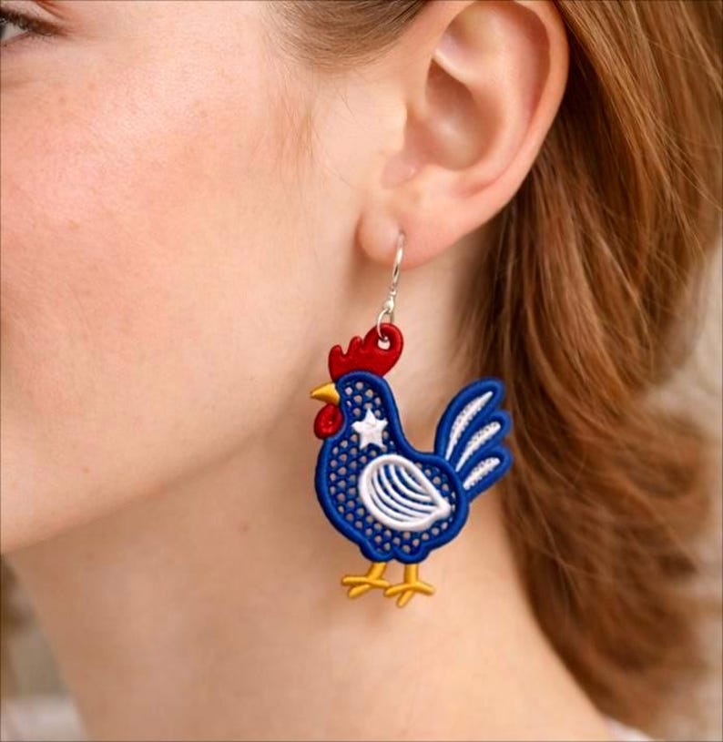 May include: A pair of rooster-shaped earrings. The earrings feature a blue body with white and red accents, including a white star. The rooster has a red comb and yellow feet. The earrings are hanging from silver hooks.