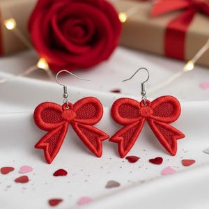 May include: A pair of red bow-shaped earrings with silver hooks. The earrings are made of embroidered fabric and are displayed on a white satin surface. A red rose and gift box are in the background.
