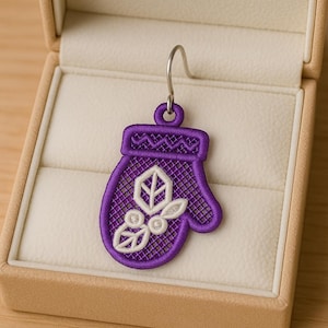 May include: A single purple mitten-shaped earring with a silver hook, displayed in a beige jewelry box. The mitten features a white embroidered design of a leaf and berries. The earring is a festive accessory.