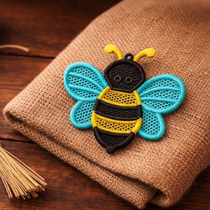 May include: An embroidered bee patch in black, yellow, and blue, resting on brown fabric. The bee design has intricate detailing. Ideal for adding to clothing or accessories, this patch is a charming decorative element.