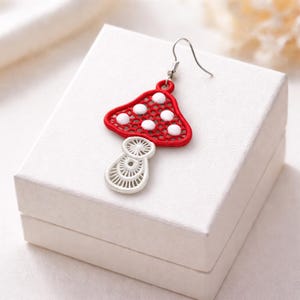 May include: A single, red and white mushroom-shaped earring with a silver hook. The mushroom cap is red with white polka dots, and the stem is white with a detailed, openwork design. The earring is set against a white background.