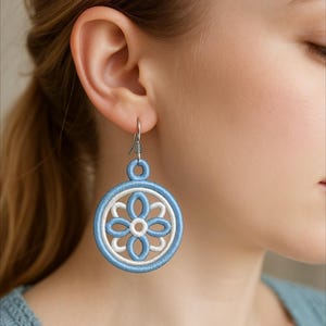 以下が含まれることがあります： 水色と白の花柄刺繍のイヤリングのクローズアップ。イヤリングは円形で、中央に花の模様があり、フック用のループが上部にあります。フックは銀色です。
