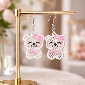May include: A pair of white bear-shaped earrings with pink bows and accents. The earrings are displayed on a gold-colored stand. The bears have black eyes and smiles, and are made of a mesh-like material.