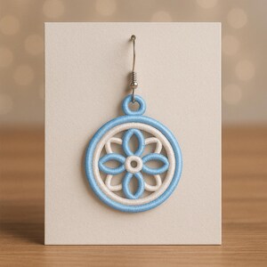 以下が含まれることがあります： 青と白の刺繍イヤリング、花の模様。 イヤリングは円形で、中央に花の模様があり、青い縁取りがあります。 銀色のフックから吊り下げられ、ニュートラルカラーのカードに表示されています。