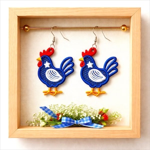 May include: A pair of rooster-shaped earrings with blue, white, red, and yellow accents. The earrings are displayed in a wooden frame with a decorative bow and floral arrangement. The rooster design features a star on the chest.