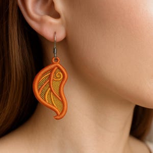 May include: An orange and yellow embroidered leaf-shaped earring with a silver hook. The earring features a detailed design with a mesh-like pattern and a swirling accent. The earring is worn on a person's ear.