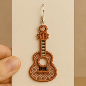 Può includere: Un orecchino a forma di chitarra marrone con un gancio argentato. L'orecchino presenta un design ricamato dettagliato, tra cui una buca e una tastiera con accenti neri e gialli. L'orecchino è tenuto su uno sfondo neutro.