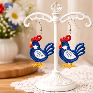May include: A pair of rooster-shaped earrings. The earrings feature a blue body with white accents, a red comb, and yellow feet. The earrings are displayed on a white decorative stand.