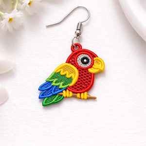 May include: A colorful parrot-shaped earring with a silver hook. The parrot is red with yellow, green, and blue accents, perched on a brown branch. The earring is likely made of fabric or embroidery.