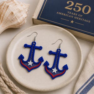 May include: A pair of anchor-shaped earrings featuring blue embroidery with a red border and a white star at the center. The earrings are constructed with a mesh-like pattern and include silver-toned metal hooks for attachment to the ear.