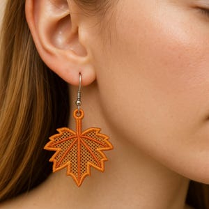 May include: Orange leaf-shaped earrings with a detailed embroidered design. The earrings are suspended from silver-colored hooks. The leaf design features a cross-hatched pattern within the outline, creating a textured appearance. The earrings are a fall-themed accessory.