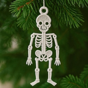 Könnte beinhalten: Weißer, gestickter Skelett-Weihnachtsschmuck. Der Schmuck hängt an einem grünen Kiefernzweig. Das Skelett hat einen Schädel, einen Brustkorb, Arme und Beine. Der Schmuck besteht aus einem gewebten Material.
