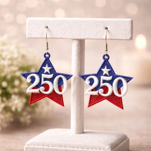 May include: A pair of star-shaped earrings in red, white, and blue. Each earring features a blue star top with a white star and the number "250" in white. The bottom of the star is red. The earrings hang from silver hooks.