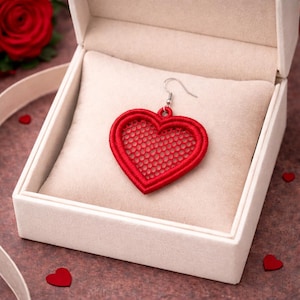 Puede incluir: Un pendiente rojo en forma de corazón con un diseño de malla delicada, exhibido en una caja de joyería blanca abierta. El pendiente tiene un gancho plateado y está colocado sobre un cojín beige. Pequeños confeti rojos en forma de corazón están esparcidos.