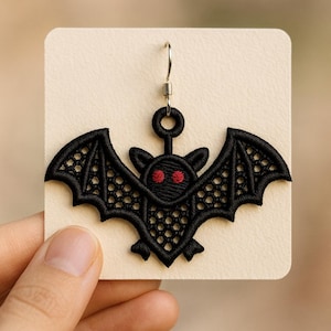 May include: Black embroidered bat earring with red eyes, displayed on a cream-colored card. The bat has detailed wing patterns and a silver earring hook. A unique, gothic-themed accessory.