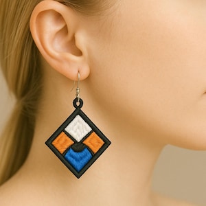 May include: Embroidered geometric earring with a black border, incorporating white, orange, and blue sections. The earring, square-shaped, has a silver hook. The design presents a modern, abstract pattern.