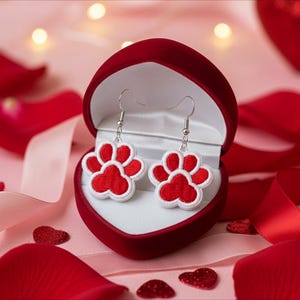 May include: A pair of red and white paw print earrings displayed in a red heart-shaped jewelry box. The earrings have a white border and silver hooks. The background features red rose petals and a satin ribbon.