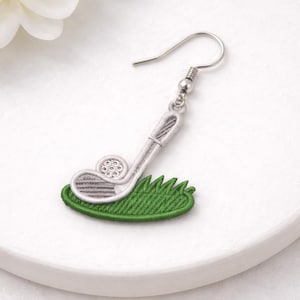May include: A silver-toned golf club earring with a detailed design, featuring a golf club over a patch of green grass. The earring has a hook closure. The design is embroidered.