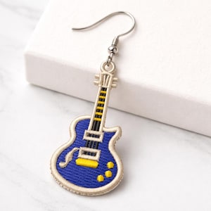Pode incluir: Um único brinco em forma de guitarra com um gancho prateado. O brinco apresenta um corpo azul com detalhes amarelos e pretos e uma borda creme. O design da guitarra é bordado.