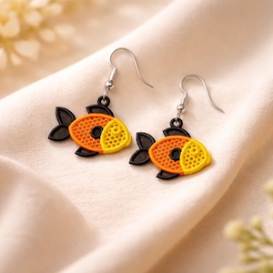 Puede incluir: Un par de pendientes con forma de pez. Los pendientes presentan cuerpos naranjas y amarillos con aletas y colas negras. Los peces tienen un diseño texturizado y están suspendidos de ganchos plateados.