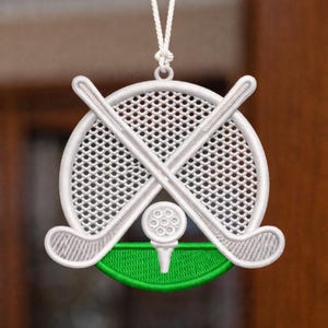 以下が含まれることがあります： 刺繍されたゴルフをテーマにしたオーナメント。交差したゴルフクラブ、ティーの上のゴルフボール、網のような背景が特徴です。デザインには緑色のベースが含まれており、白い紐で吊り下げられています。オーナメントは白と緑のアクセントです。