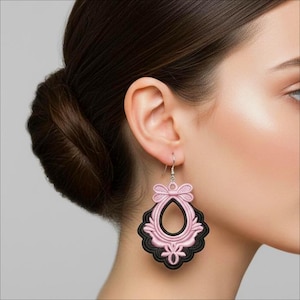 May include: A close-up of a pink and black filigree earring. The earring has a teardrop shape with a bow at the top and a decorative black border. The earring is suspended from a silver hook.