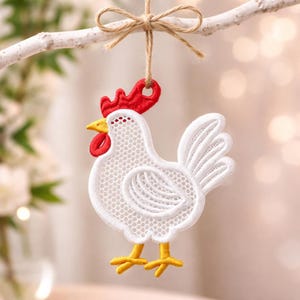 Könnte beinhalten: Ein weißes Spitzen-Hahn-Ornament mit rotem Kamm und Kehllappen, gelben Füßen und gelbem Schnabel. Der Hahn hängt an einer Schnurschleife, die an einem weißen Ast befestigt ist. Das Ornament ist ein dekorativer Gegenstand.