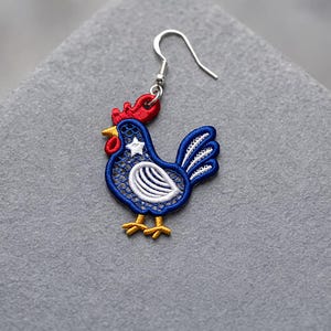 May include: A single rooster-shaped earring. The earring features a blue body with white and red accents, including a star and a comb. The rooster has a silver hook for easy wear. The design is detailed and colorful.