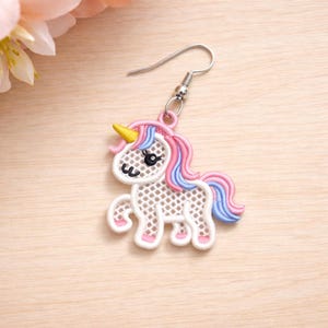 Könnte beinhalten: Ein verspielter Einhorn-Ohrring mit einem weißen, durchbrochenen Körper, einer rosa und blauen Mähne und einem Schweif sowie einem gelben Horn. Der Ohrring hat einen silberfarbenen Haken und kleine rosa Akzente an den Füßen. Ein niedliches, fantasievolles Accessoire.
