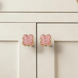 Op de afbeelding: Vier kastknoppen met een klavervormig ontwerp. De knoppen hebben een roze, gemarmerd midden en een goudkleurige rand. Ze zijn gemonteerd op een witte kast. Op de achtergrond staan een paar bruine en oranje orchideeën.