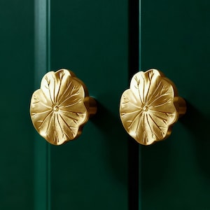 May include: Two gold-coloured, leaf-shaped cabinet knobs are mounted on a dark green cabinet door. The knobs have detailed veining and a central circular design. The background is a solid, dark green colour.