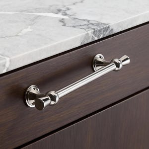 Può includere: Una maniglia per armadio cromata lucida con una barra cilindrica e terminali decorativi. La maniglia è montata su un'anta di un armadio marrone scuro, con un piano di lavoro in marmo sullo sfondo.