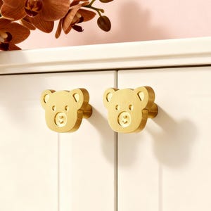 Op de afbeelding: Twee goudkleurige kastknoppen in de vorm van teddybeergezichten. De knoppen zijn gemonteerd op een witte kastdeur. Op de achtergrond een tak met bruine orchideeën.