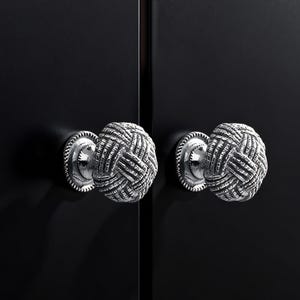 Pode incluir: Close-up de dois puxadores de armário prateados com um design de nó tecido. Os puxadores são montados numa porta de armário preta. Os puxadores têm uma borda decorativa em forma de corda.
