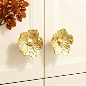 May include: Two gold-colored, flower-shaped cabinet knobs. The knobs are mounted on a white cabinet door. The flower knobs have detailed petals and a textured center. A branch of brown orchids is in the upper left corner.