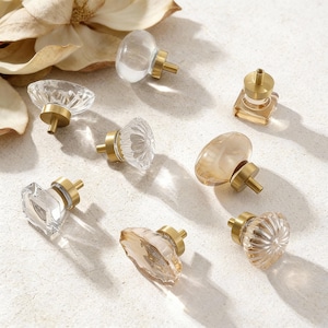 May include: A selection of decorative cabinet knobs in different shapes and colours. The knobs have clear and amber-toned glass or crystal elements with brass-coloured metal bases. The shapes include oval, square, and round designs, offering a range of styles.