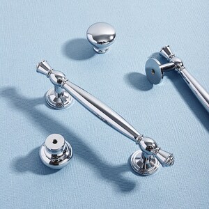 May include: A collection of polished chrome cabinet hardware, including two bar pulls, a cabinet knob, and a smaller knob. The hardware has a reflective surface and is arranged on a light blue surface.